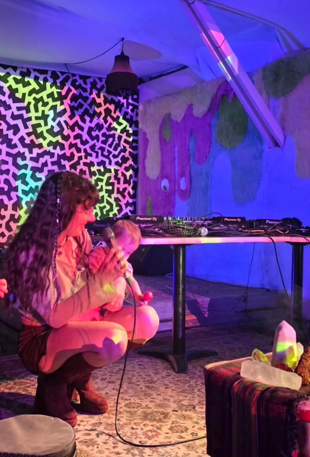 Mother Mystica holding a microphone while sitting with a young child during a colorful live gathering